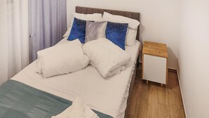 Una cama bien hecha con sábanas blancas y almohadas azules, una mesita de noche pequeña y un suelo de madera.