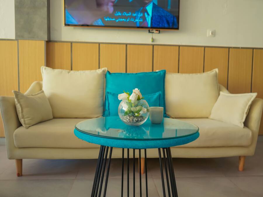 A beige sofa with a glass coffee table in front, a teal pillow, and a vase with white flowers.