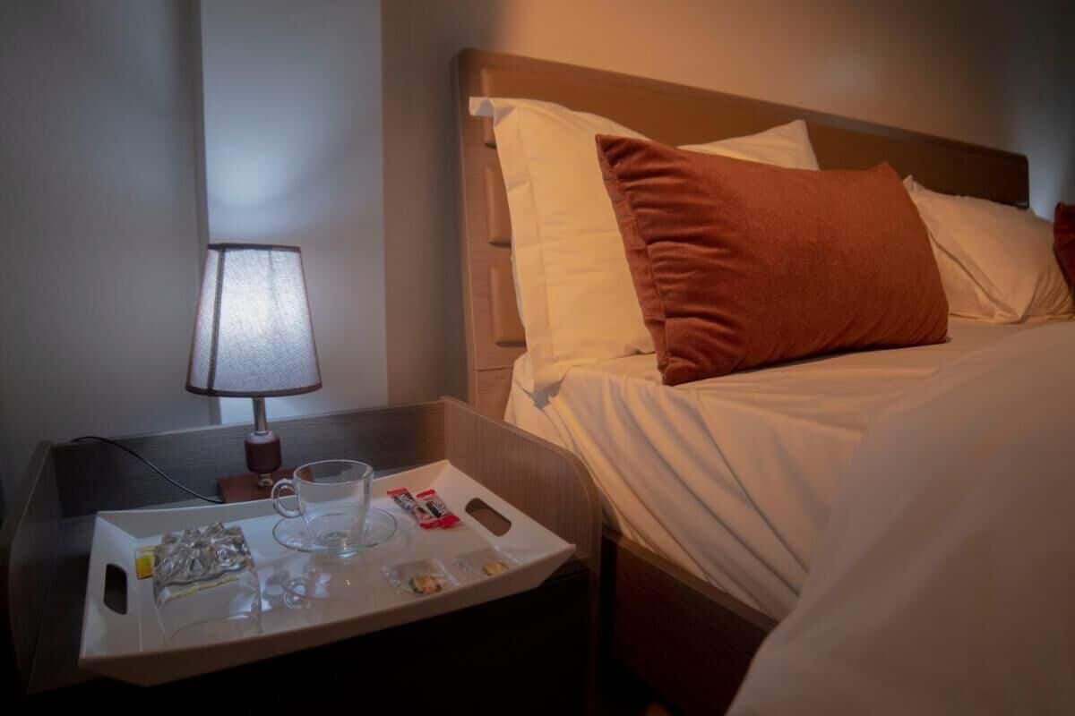 Habitación de hotel con una cama bien hecha, una mesita de noche con una lámpara y una bandeja con agua y aperitivos.
