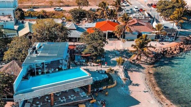 An aerial view of a beachside resort with a large building featuring a blue roof, a pool area, and a sandy beach. - Beautiful condo in La romana, DR with pool and walking distance to Caleta beach. (La Romana)