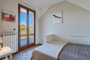 A bedroom with a bed, a radiator, a table with a lamp, a door leading to a balcony, and a wall with a picture of a van and a Dolce Vita sign.