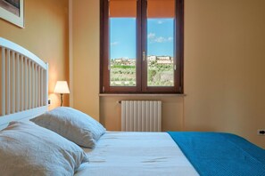 Un dormitorio con cama, ventana con vistas a edificios, radiador y lámpara.