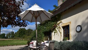 A patio with a table and chairs under a white umbrella, a clock on the wall, and a garden with gravel and greenery. - Holiday Home in Le Châtelet with Pool (Le Châtelet)