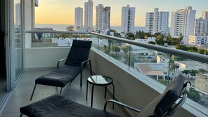Ein Balkon mit zwei schwarzen Liegestühlen und einem kleinen Tisch, mit Blick auf eine Stadtlandschaft aus hohen Gebäuden und eine Meeransicht.