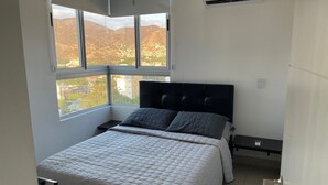 Ein Schlafzimmer mit einem großen Fenster, einem Bett mit gesteppter Bettdecke, einem schwarzen, gepolsterten Kopfteil und einer Klimaanlage an der Wand.