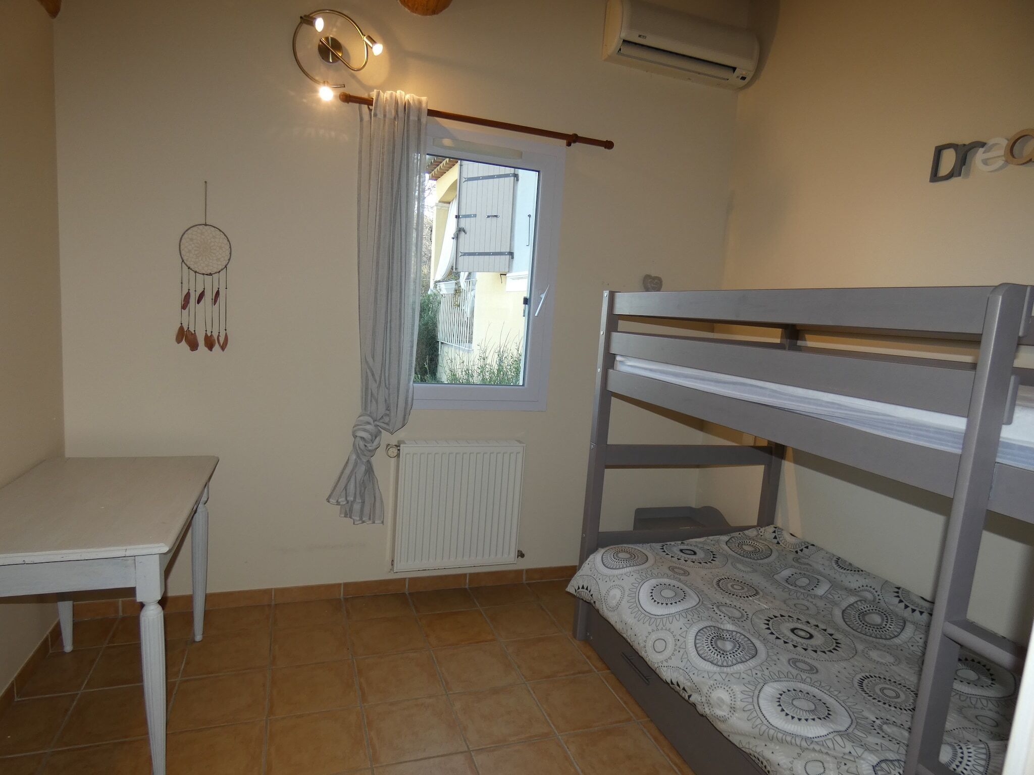 Ein kleiner, ordentlicher Raum mit Etagenbett, einem Tisch, einer Heizung und einem Traumfänger an der Wand.