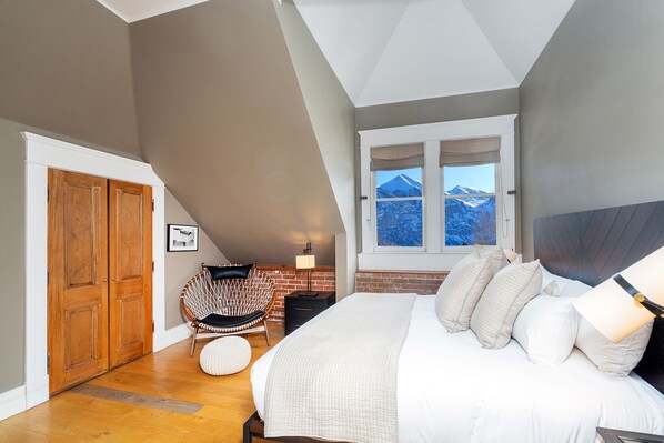 A bedroom with a large bed, a window with a mountain view, a wooden wardrobe, and a wicker chair. - Historic Building w/ Majestic Top Floor Views (Telluride)