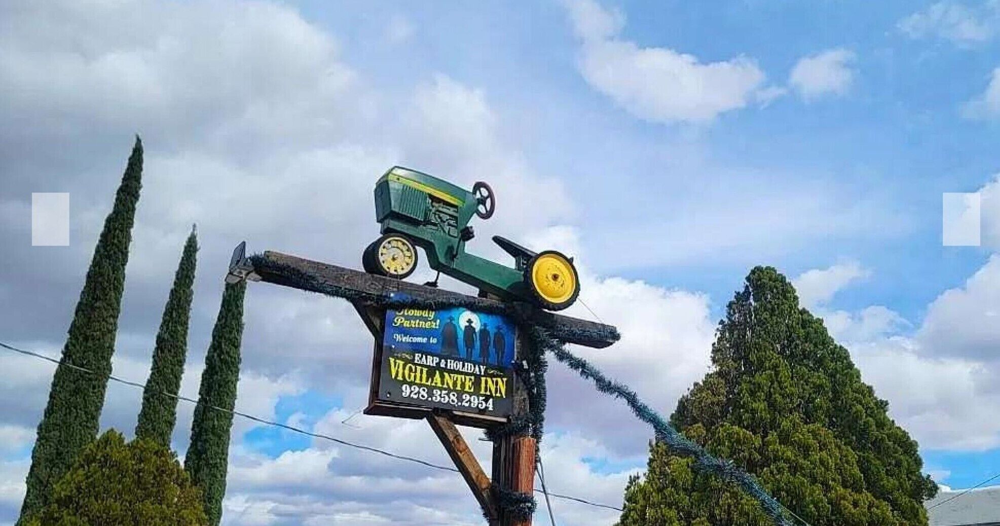 A sign for Vigilante Inn with a tractor and a clock, surrounded by tall trees.