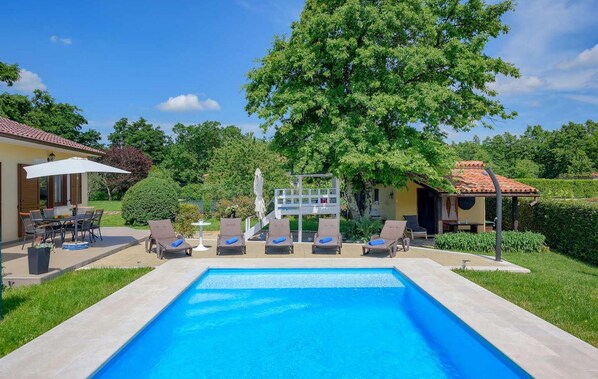 Ein Pool mit Liegestühlen und einem Tisch, umgeben von Grünpflanzen und einem Gebäude mit einem Ziegeldach.