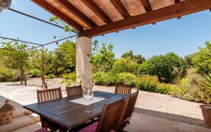 An outdoor patio with a wooden table and chairs, surrounded by greenery and a clear sky.