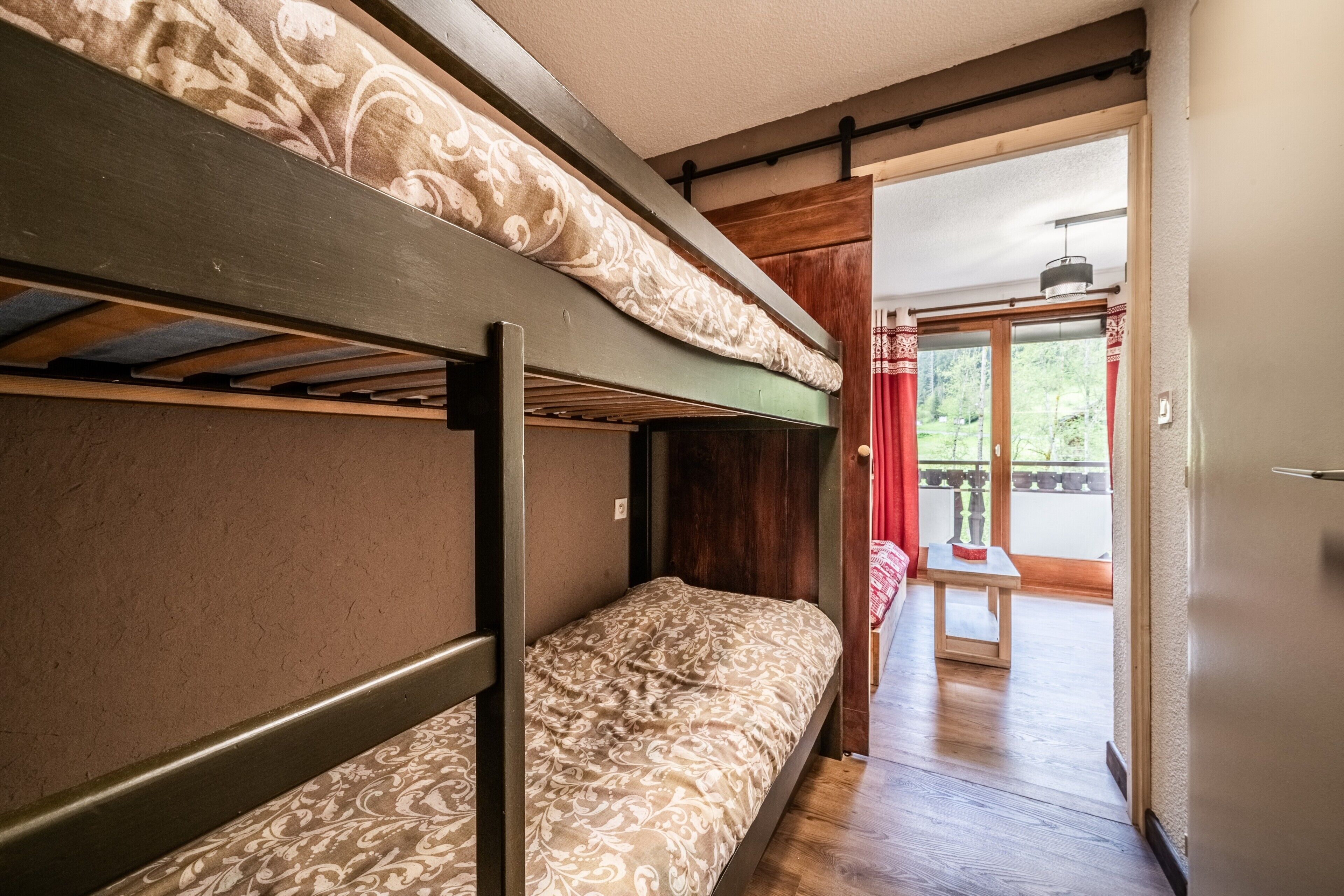 A bunk bed room with a wooden ceiling, a small table, and a view of a garden through a glass door.