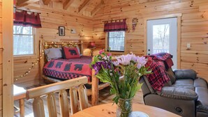 A cozy cabin interior with a wooden interior, a bed with a red and black blanket, a leather sofa, and a table with a vase of flowers. - Bear Hugs cabin nestled in the woods. (Logan)