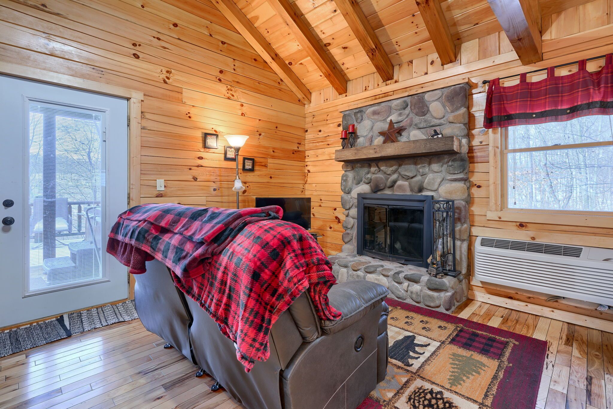 A cozy cabin interior with a stone fireplace, a reclining chair with a blanket, wooden walls, and a large window.