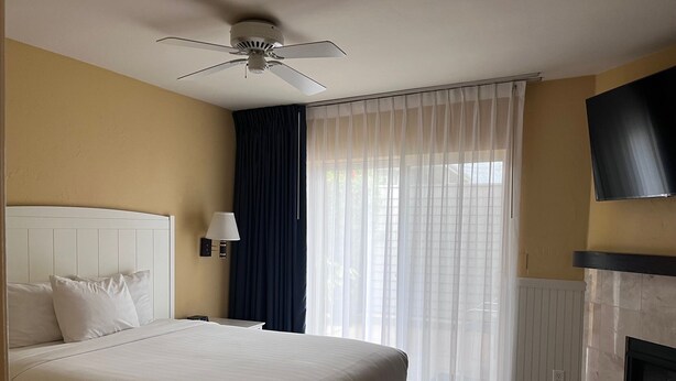 A hotel room with a bed, a ceiling fan, a wall-mounted TV, a fireplace, and sheer curtains.