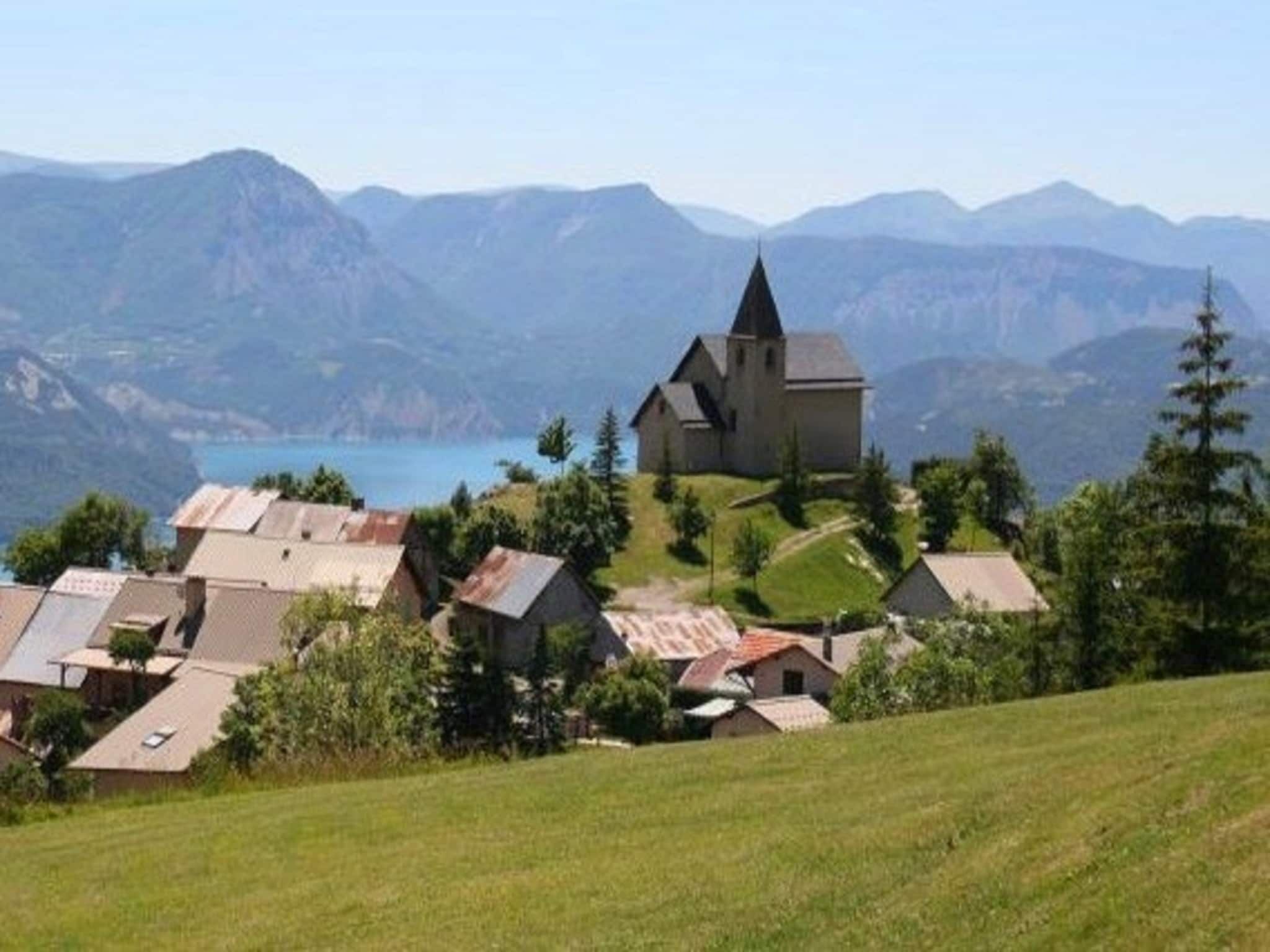 A church with a steep roof and a bell tower, surrounded by houses with sloped roofs, set against a backdrop of mountains and a lake.