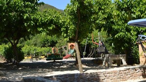 Un parque infantil con diversas estructuras de juego, rodeado de árboles y un muro de piedra.