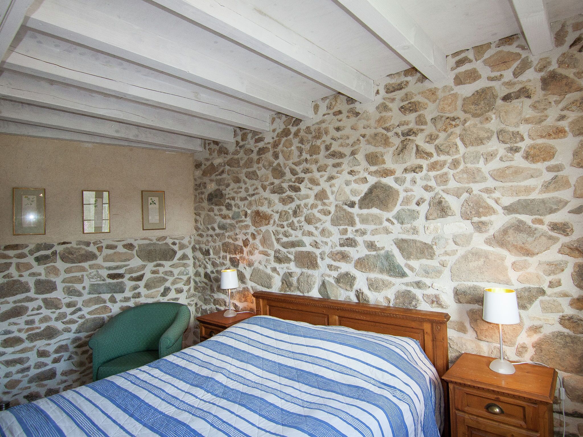 Un dormitorio con pared de piedra, una cama con colcha a rayas, una silla verde y dos mesitas de noche con lámparas.