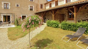 A building with a stone facade, a covered patio area, and a small garden with a flower bush and two lounge chairs.
