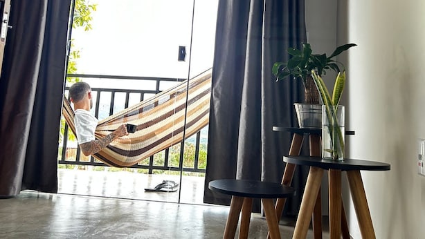 A room with a large sliding glass door, a hammock, a small table with a plant, and a wooden floor. - Flat Aconchegante de Frente Para as Montanhas e Rodeado Pela Natureza (Alto Paraíso de Goiás)