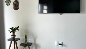 A modern living room with a flat-screen TV mounted on the wall, two small round tables with potted plants, and a decorative Buddha head sculpture. - Flat Aconchegante de Frente Para as Montanhas e Rodeado Pela Natureza (Alto Paraíso de Goiás)