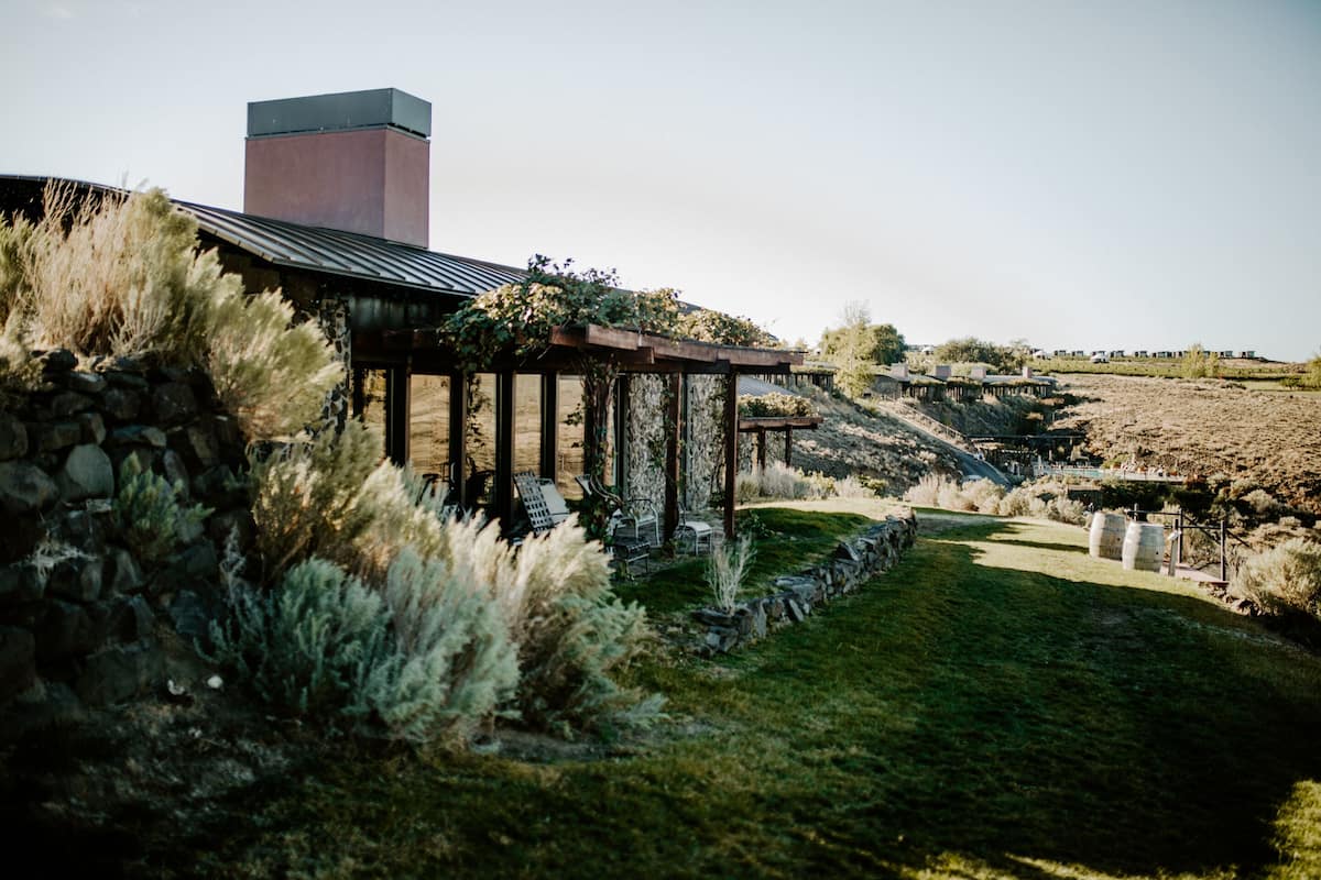 A stone wall with a pergola and seating area, a stone chimney, and a grassy landscape with distant buildings.