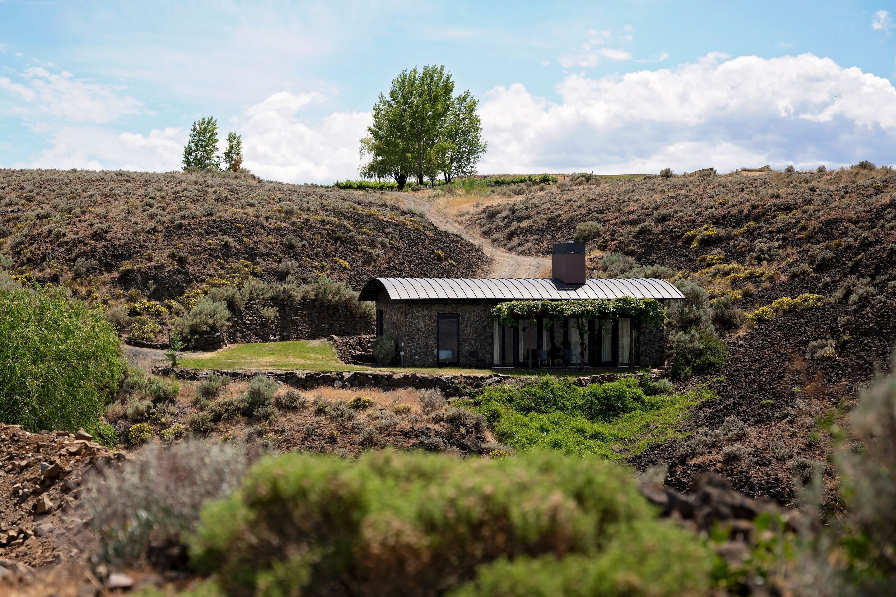 A rustic stone house with a metal roof, surrounded by dry vegetation and a clear sky.
