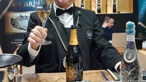 Un homme en costume noir, tenant un verre à vin, est assis à une table sur laquelle se trouvent une bouteille de vin, une assiette décorative et une bouteille emballée dans du plastique.