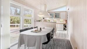 A dining area with a table set for four, large windows, a kitchen with white cabinets, and a ceiling with wooden beams.