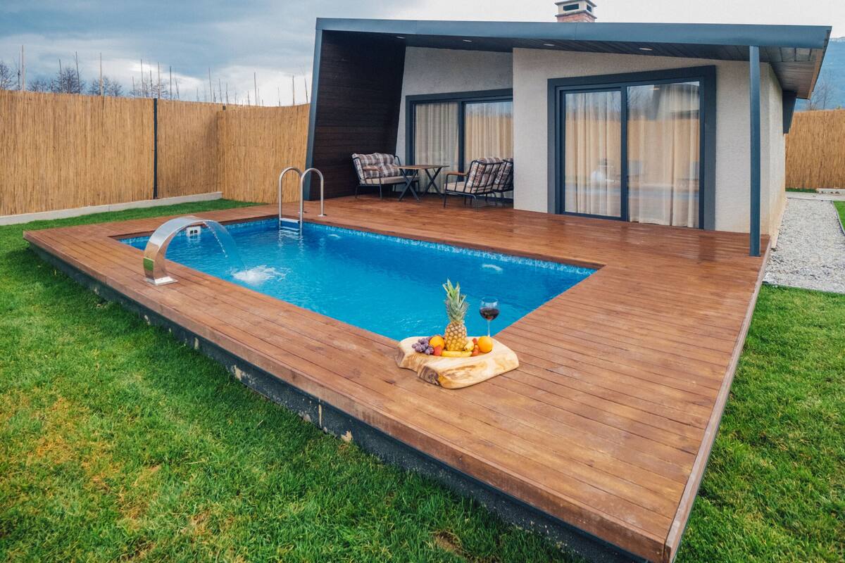 A modern outdoor pool area with a wooden deck, a lounge chair, and a table with fruits and a glass of wine.