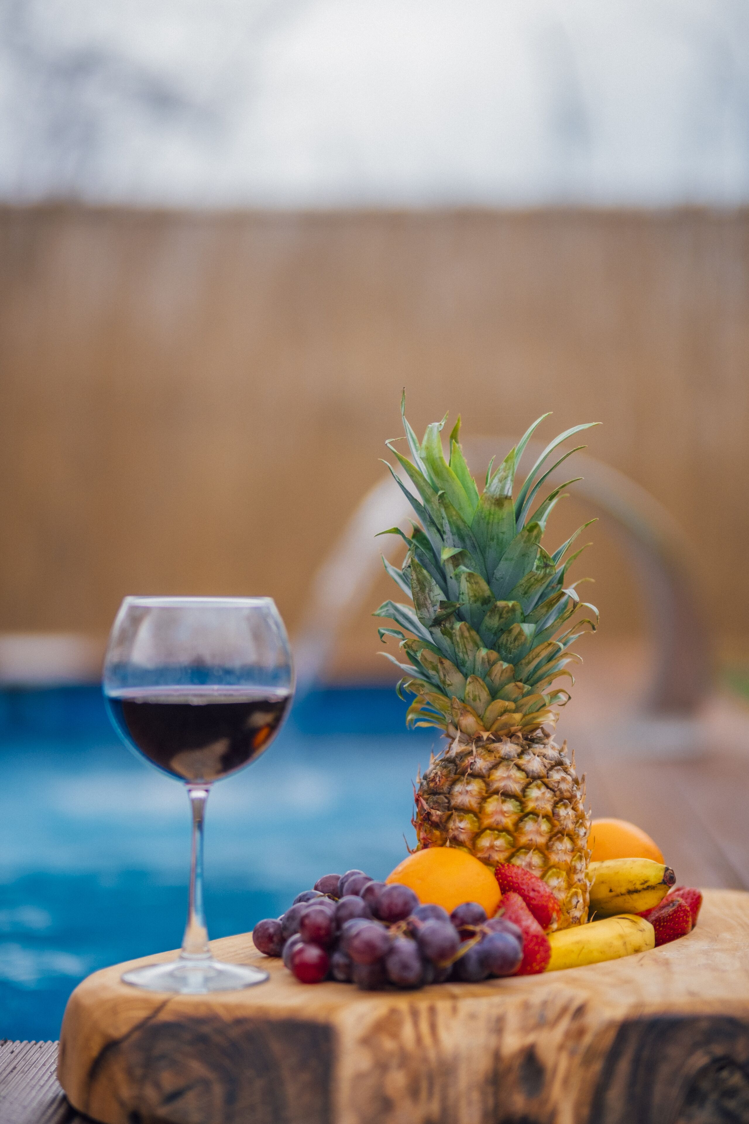 A wooden table with a glass of red wine, a pineapple, grapes, a banana, and an orange.