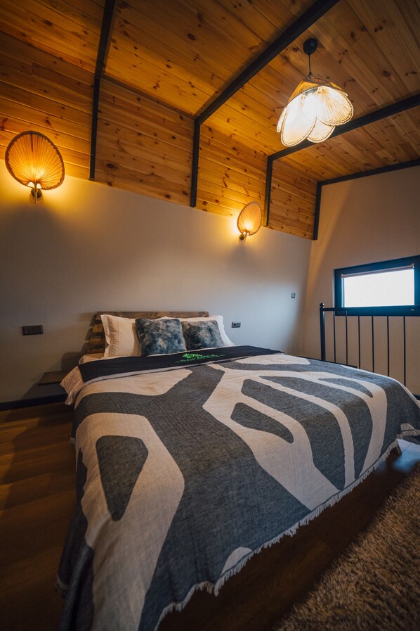 A bedroom with a wooden ceiling, a bed with a patterned blanket, two wall-mounted lamps, and a window.