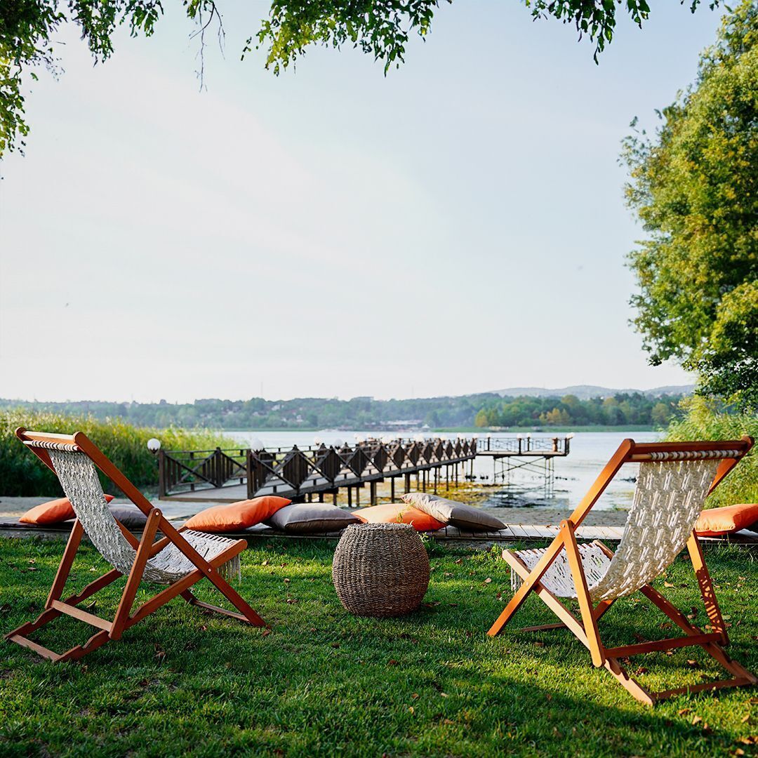 A lakeside seating area with two wooden chairs and a woven ottoman.