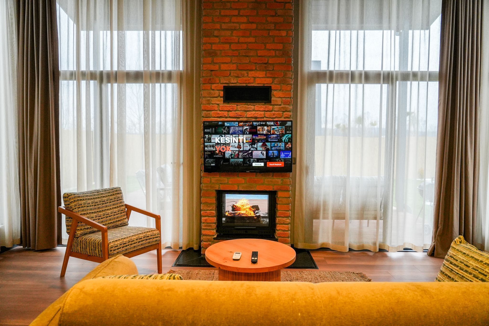 A living room with a brick fireplace, a flat-screen TV, a wooden armchair, and a yellow sofa.