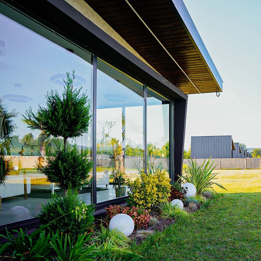 A modern house with large glass windows, a wooden roof, and a well-maintained garden with various plants and spherical decorations.