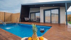 A wooden deck with a pool, a table with fruit and a glass of wine, and a modern building in the background. - Vital Göl (Sapanca)