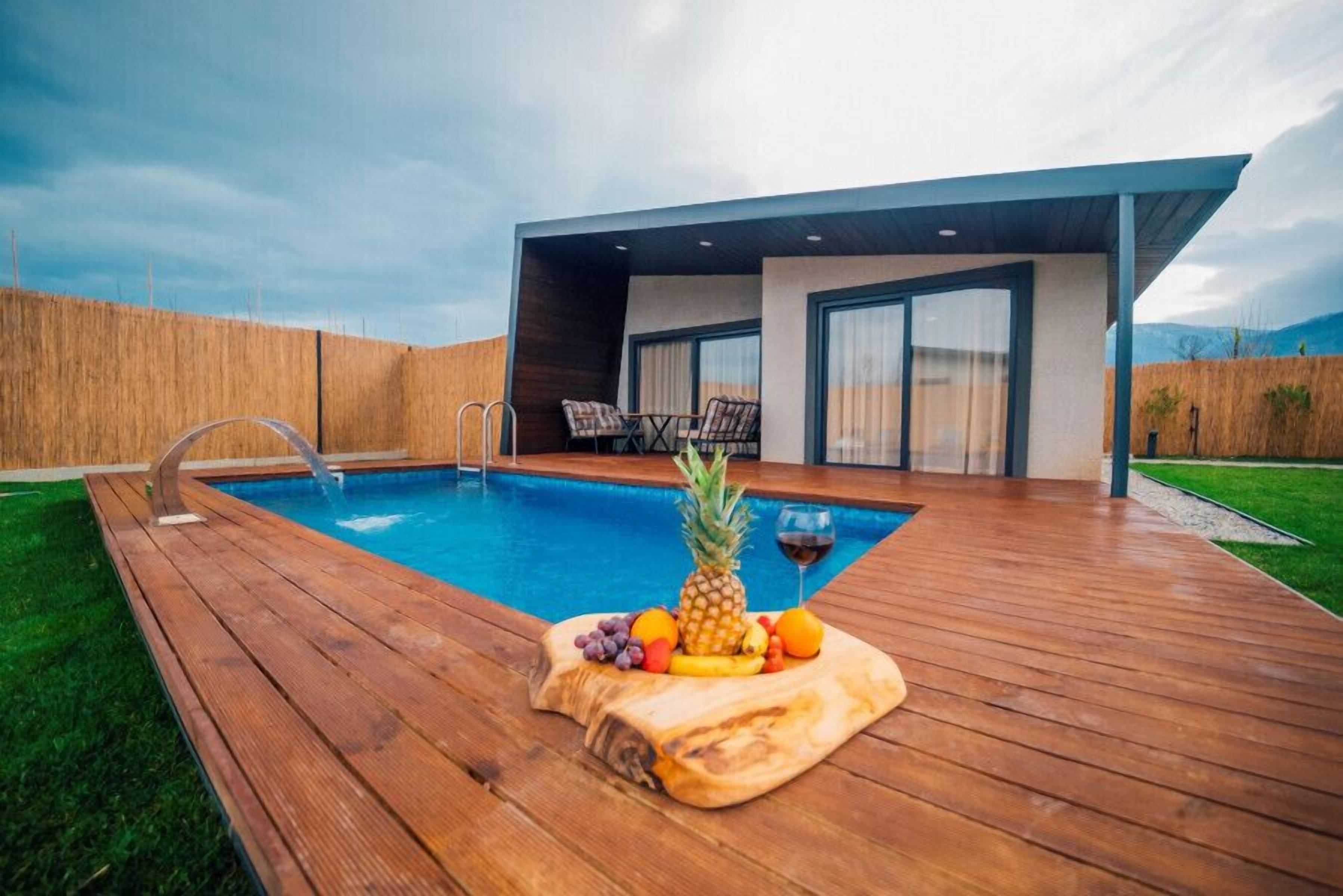 Un área de piscina moderna al aire libre con cubierta de madera, una presentación de frutas tropicales y un espacio de estar con sillas y una mesa.