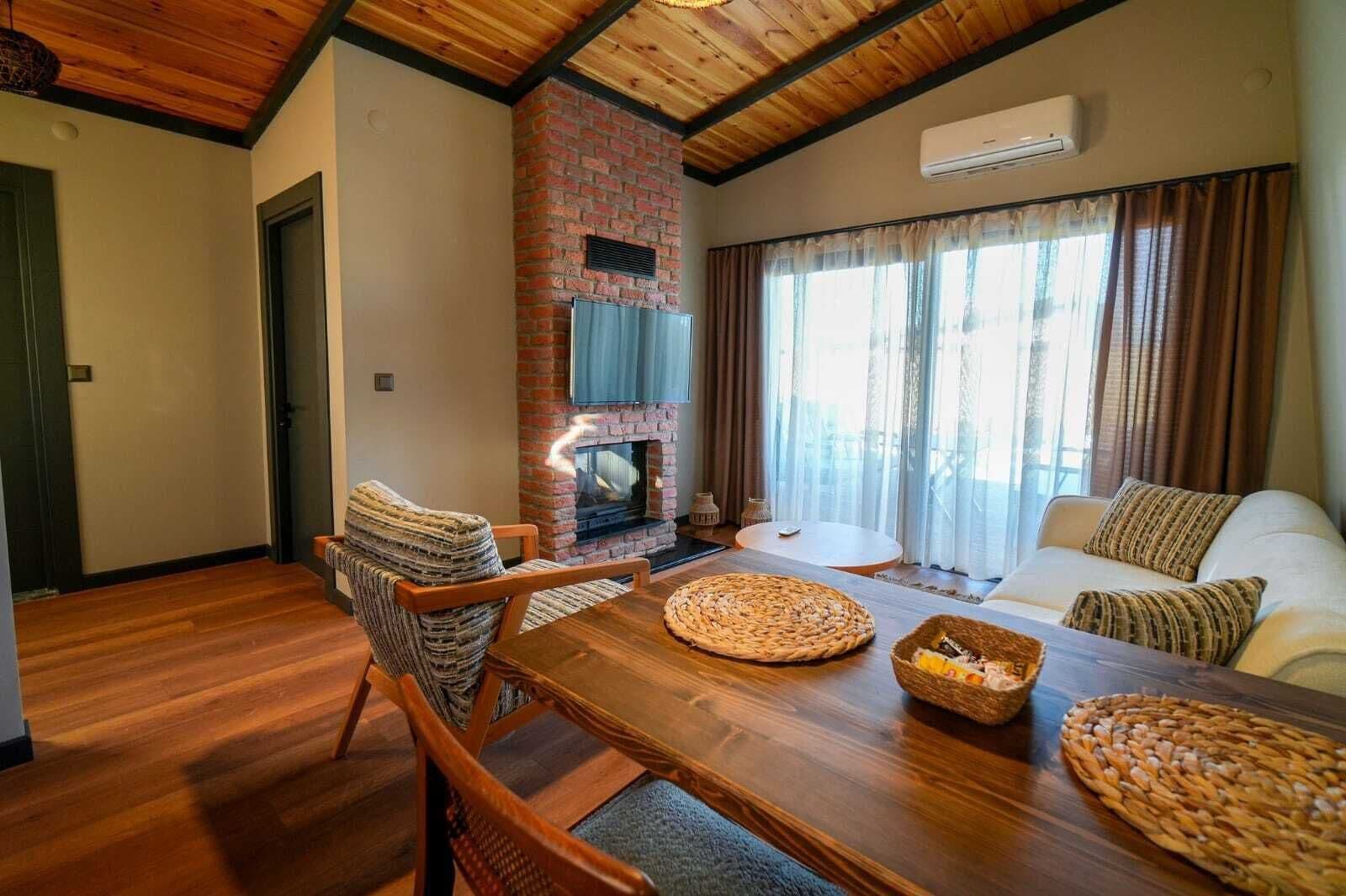 A cozy living room with a brick fireplace, wooden furniture, and a large window with curtains.