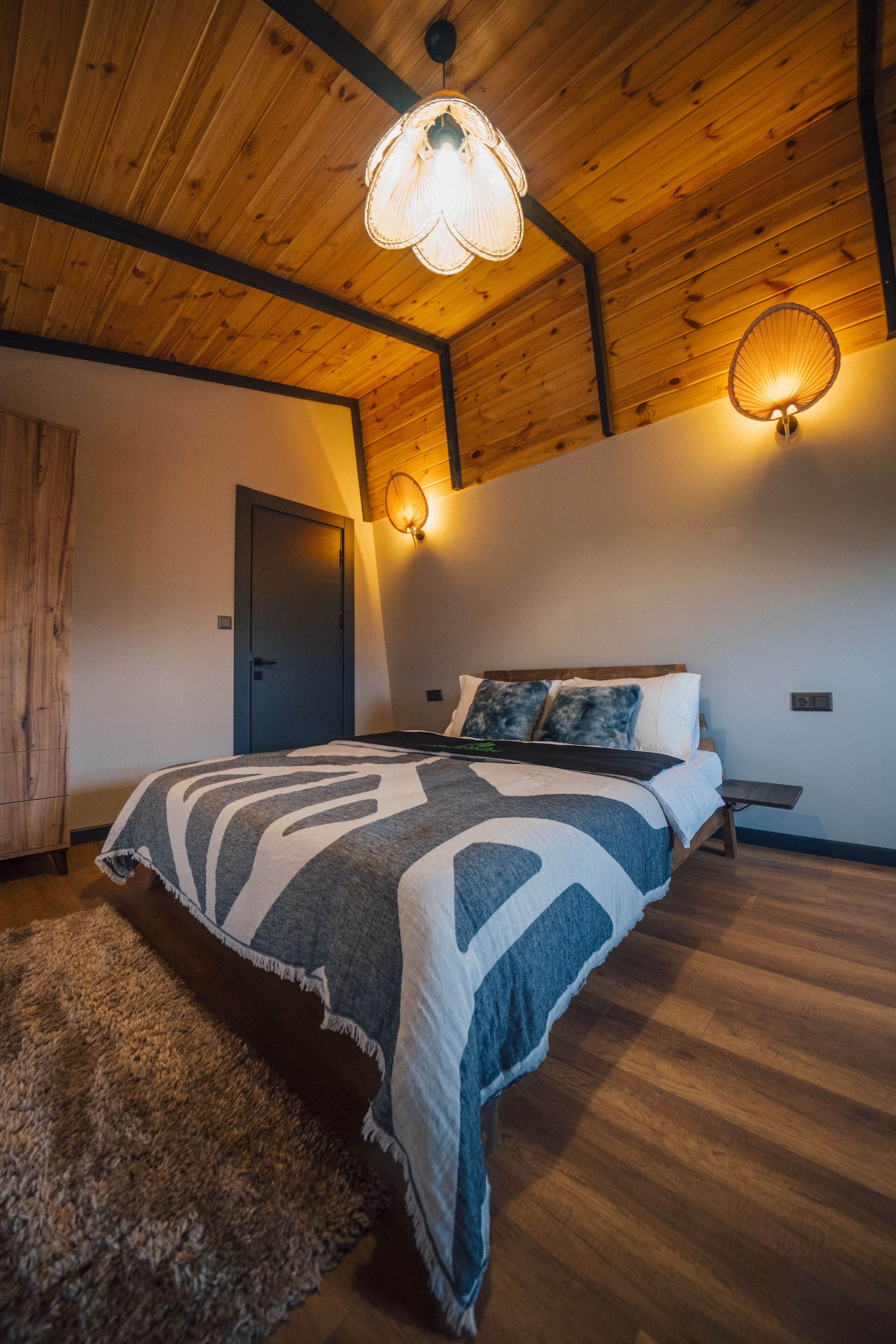 Een slaapkamer met een houten plafond, een bed met een geborreld deken, twee wandlampen en een deur.