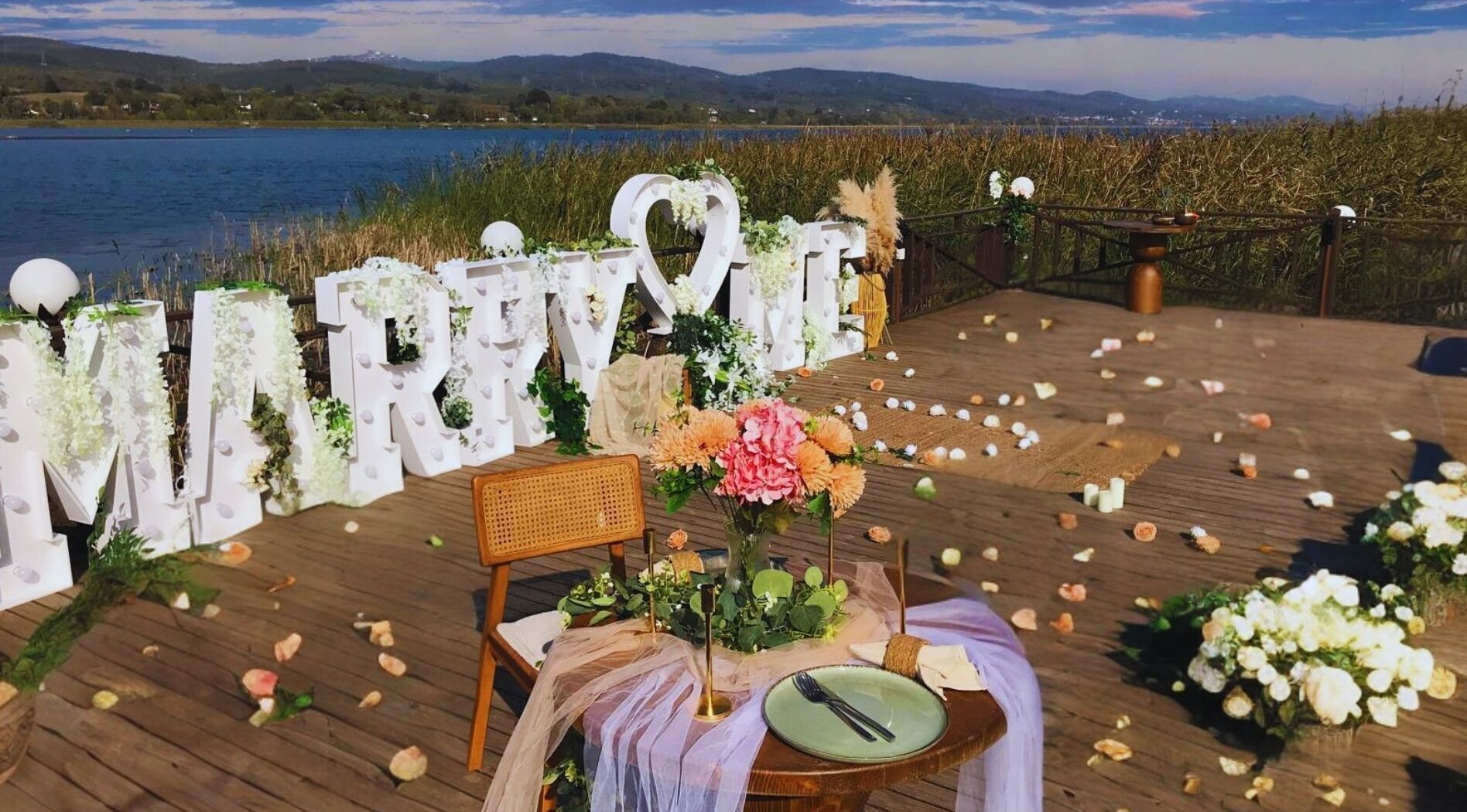 A wedding setup with a wooden deck, a chair, a table with a green plate, and a backdrop with 'MARRY ME' letters.