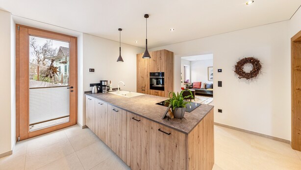 Een moderne keuken met houten kastjes, een centraal eiland, hanglampen en uitzicht door een schuifdeur.