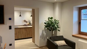 Een moderne woonkamer met een donkere bank, een potplant en een keuken op de achtergrond.