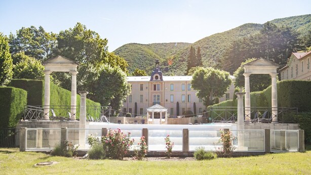 Um edifício com um pavilhão central, rodeado por um jardim aparado e árvores.