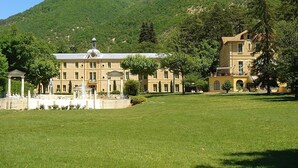 Un gran edificio de estilo clásico con una fuente en primer plano, rodeado de un césped cuidado y árboles, con colinas verdes al fondo.