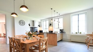 Una cocina moderna con una isla central, una mesa de comedor de madera y sillas. Se observan luces colgantes, un reloj y ventanas amplias que dejan entrar luz natural.