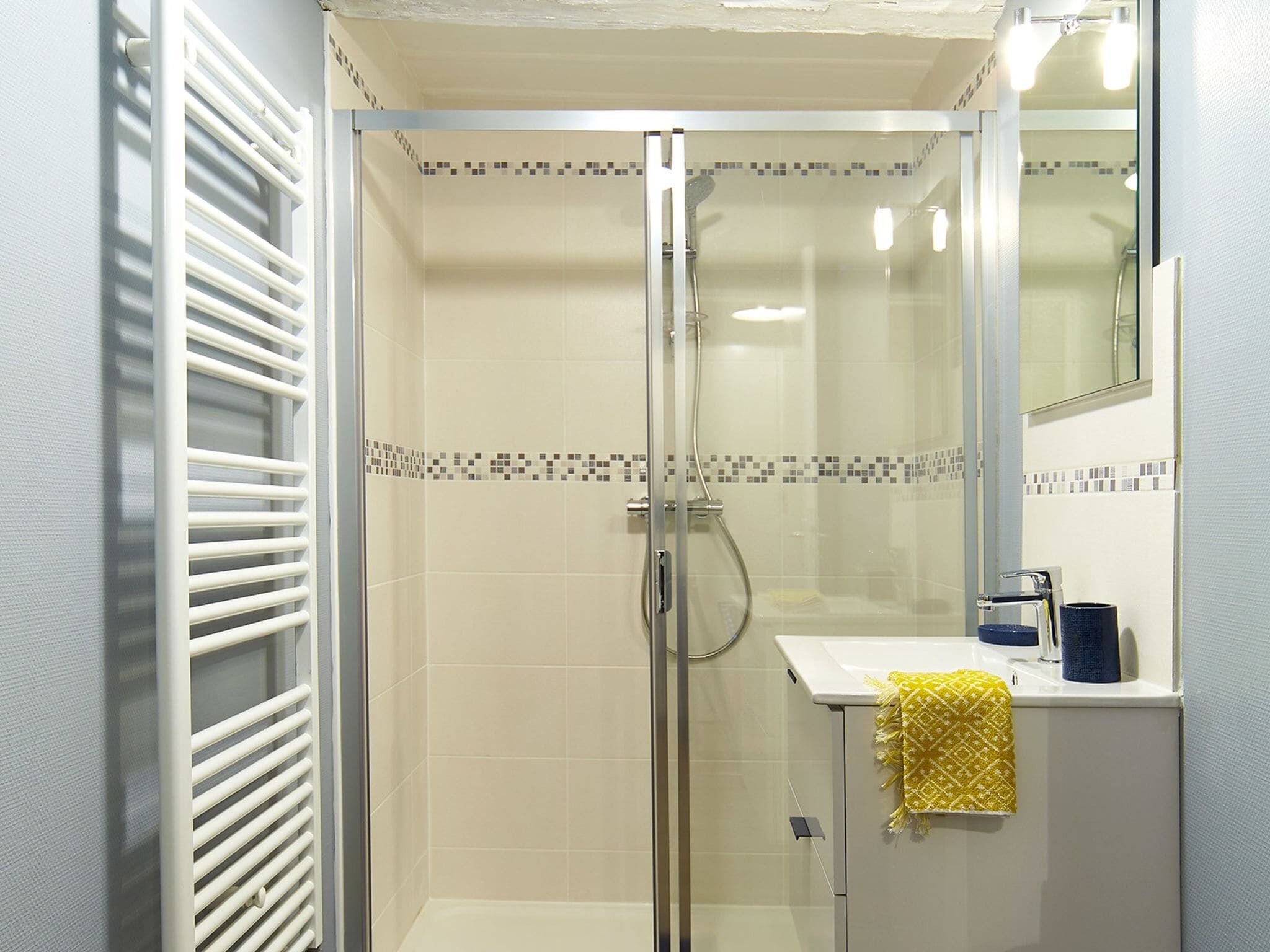 Une douche fermée par des parois de verre, avec un mur recouvert de carreaux blancs, une porte en verre et un petit lavabo agrémenté d’une serviette jaune.