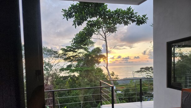 A modern balcony with a view of the sunset over a body of water, surrounded by lush greenery. - 1bdr Stunning House Uvita CUSINGA | Panoramic Ocean View (Bahía Ballena)