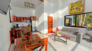 A modern living room with a wooden dining table, a grey sofa, and a coffee table. There is a kitchen area with wooden cabinets and a refrigerator. - 1bdr Stunning House Uvita CUSINGA | Panoramic Ocean View (Bahía Ballena)