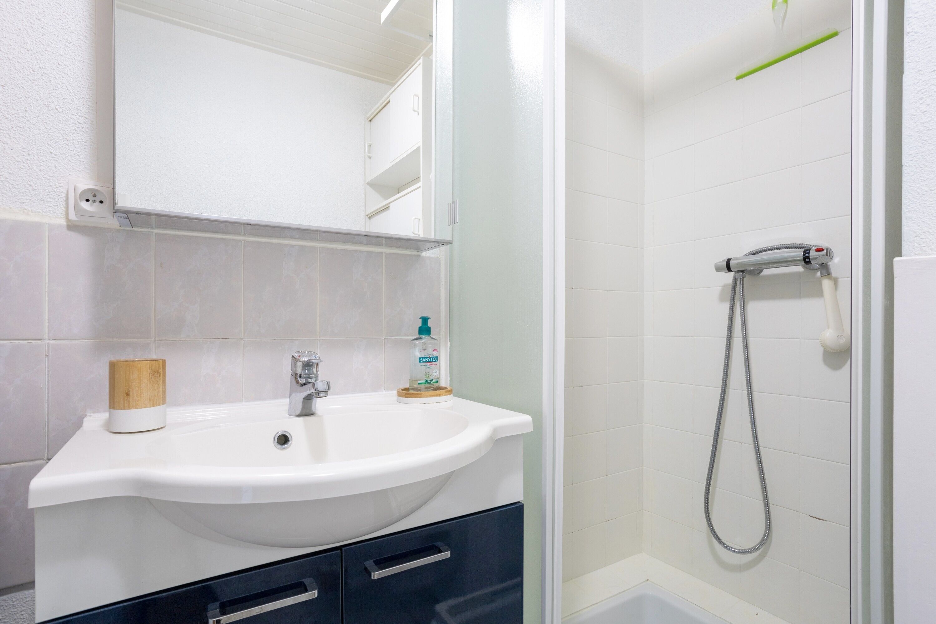 A bathroom with a white sink, a mirror, a shower, and a wooden cup holder.