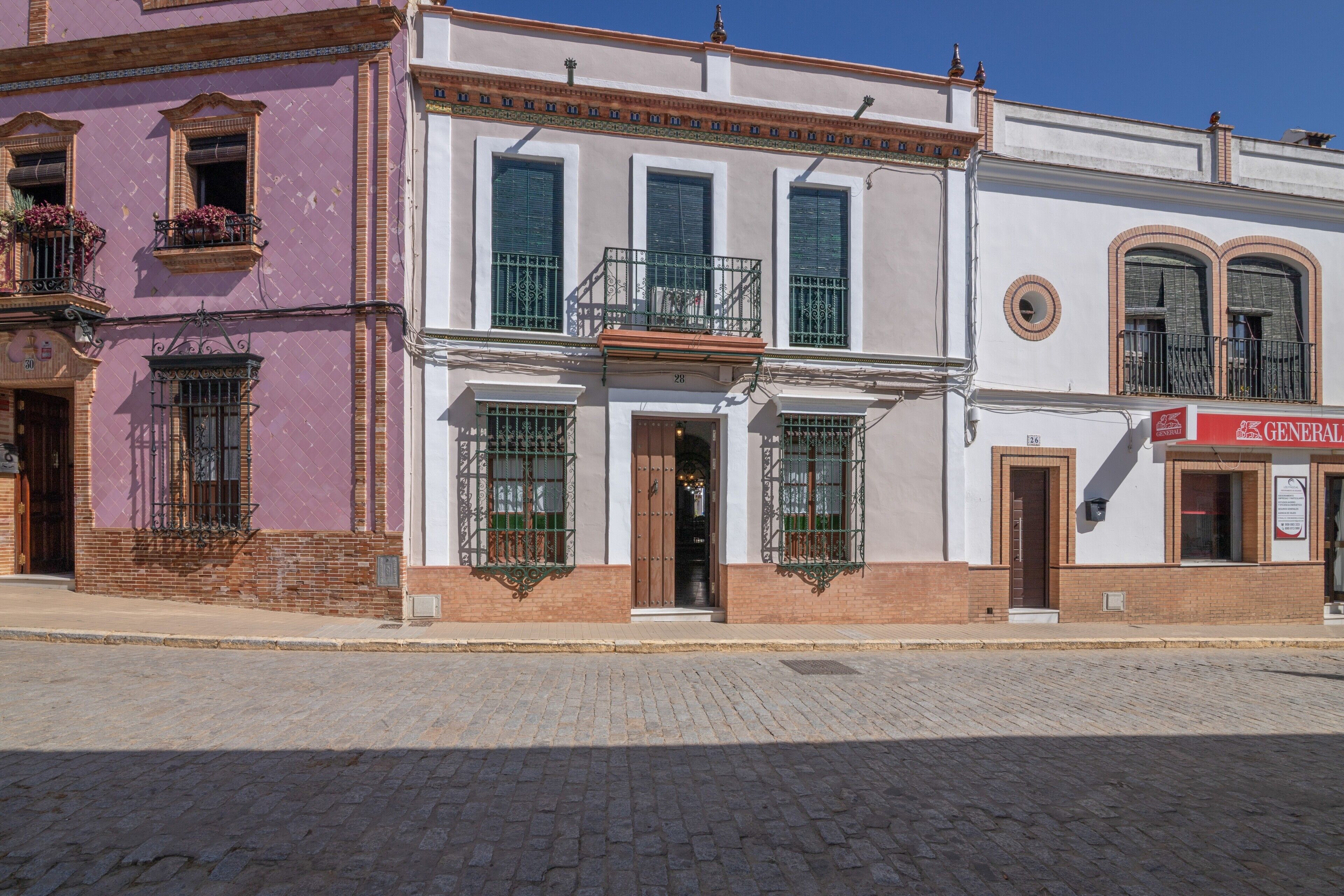 Vista de la calle con un edificio que tiene una entrada con puertas dobles, un balcón con barandas de metal y un letrero de una tienda de abarrotes.