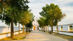 Un chemin menant à un bâtiment doté d’arcades et d’un clocher, entouré d’arbres et d’un mur blanc.